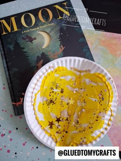 A book titled “Moon” sits beside a paper plate moon covered in yellow paint and glitter, suggesting a creative, paint splat moon-themed craft activity. The handles @GLUEDTOMYCRAFTS are visible on the image.