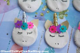 Handmade clay unicorn magnets crafted from white clay, featuring closed eyes, gold or silver paper horns, gold ears, and colorful flower decorations. The crafts are displayed on a pastel background.