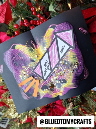 A colorful, abstract New Year’s craft made with marbled paint fireworks, glitter, and paper cutouts is displayed in front of a Christmas tree. “Happy New Year 2024” is written inside a triangle. Username @GLUEDTOMYCRAFTS appears at the bottom.