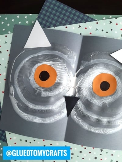 A paint splat owl face craft made from white paint swirled on gray paper, with orange and black paper eyes, a black paper beak, and pointed white ears. Patterned papers are underneath. @GLUETOMYCRAFTS appears in a blue box.
