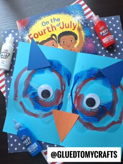 Bright blue and red paper owl craft with a fun paint splat owl face and large googly eyes sits on a patriotic background, surrounded by bottles of puffy paint, with a Fourth of July-themed book in the background. @GLUEDTOMYCRAFTS watermark at bottom.