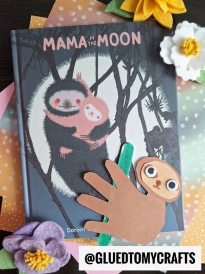 A children’s book titled “Mama in the Moon” features an illustration of two sloths on the cover. Next to it is a charming handprint sloth craft, made from a traced handprint, felt flowers, and the handle of a green craft stick.