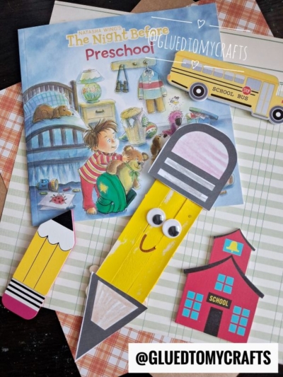A children’s book, paper pencils, a school bus, and a red schoolhouse made from craft materials are arranged on plaid paper backgrounds. A charming paint splat owl face adds a whimsical touch to the scene. The username @GLUEDTOMYCRAFTS is visible on the image.