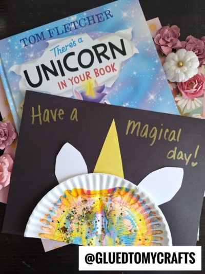 A children’s book titled “There’s a Unicorn in Your Book” sits next to a paper plate unicorn craft with paint splat unicorn hair, white and gold paper, and glitter, with “Have a magical day!” written on black paper.