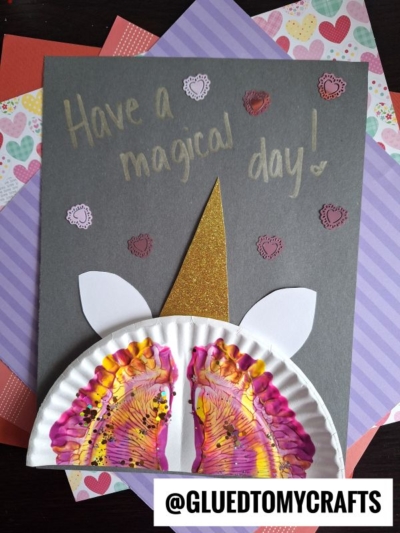 A handmade paint splatter unicorn card with a gold horn, white ears, and a paper plate decorated with colorful painted footprints for the snout, set on heart and striped paper. Text says, Have a magical day!.