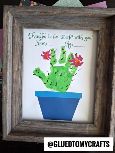 A framed cactus handprint art of a green handprint cactus in a blue pot, decorated with pink flowers and black spines. Text above reads, Thankful to be stuck with you! Name ____ Age ____. @GLUEDTOMYCRAFTS is at the bottom.