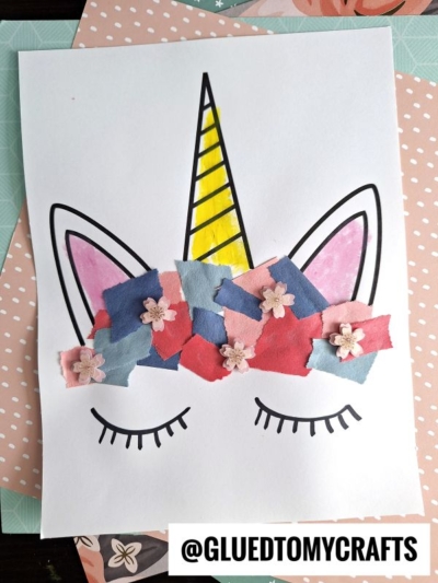 A torn paper unicorn drawing with closed eyes, a yellow horn, pink ears, and a mane crafted from blue, pink, and red paper bits with tiny flower accents on a white sheet. @GLUEDTOMYCRAFTS is at the bottom.