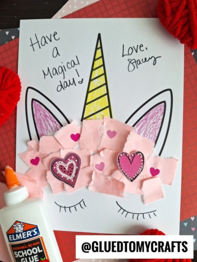 A torn paper unicorn face craft made with colored paper, pink torn paper for the muzzle, and heart decorations. Text says Have a magical day! Love, Stacey. Elmers glue and red fabric are beside the paper.
