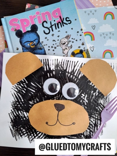A children’s book titled Spring Stinks lies behind a fork painted bear craft made with black paint, brown paper shapes, large googly eyes, and a plastic fork. The Instagram handle @GLUEDTOMYCRAFTS is at the bottom.
