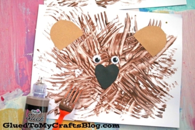 A charming fork painted bear face made from brown painted fork strokes, with googly eyes, paper ears, and a black heart nose on white paper. A fork and brown paint bottle are nearby.