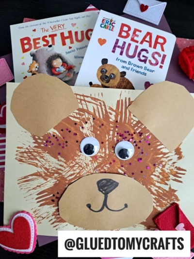 A fork painted bear craft made from brown paint, googly eyes, and paper ears and nose sits on a table with heart decorations and two bear-themed children’s books, “The Very Best Hug” and “Bear Hugs!”.