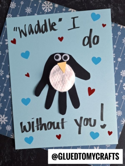 A handmade penguin handprint card featuring a black paper handprint penguin, white belly, and yellow beak. The card says "Waddle I do without you!" and is decorated with red and blue hearts—a perfect penguin keepsake craft.