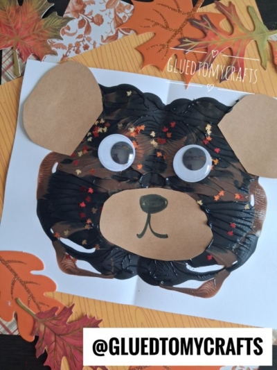 A paint splat teddy bear face craft made from painted paper with googly eyes, brown paper ears, and a snout, decorated with leaf confetti, sits on a background of autumn-themed leaves.