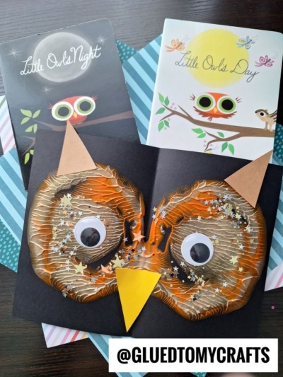 A handmade paint splat owl face card with big googly eyes, a yellow beak, and star confetti sits in front of two “Little Owls” books. Striped and polka dot paper is underneath. Text: @GLUEDTOMYCRAFTS.