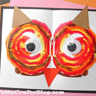 A colorful paint splat owl face on white paper features large googly eyes, brown paper ears, and an orange beak, displayed on a pink background.