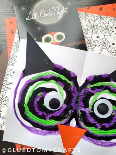 A children’s book titled Little Owl’s Night and a colorful paint splat owl face craft with googly eyes, black ears, and an orange beak on top of Halloween-themed papers.