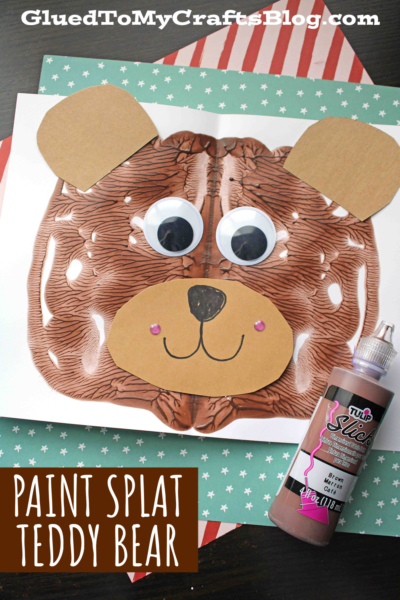 A paint splat teddy bear craft with googly eyes, a paper nose, ears, and mouth sits on patterned paper beside a bottle of brown fabric paint. Text reads “Paint Splat Teddy Bear.”.