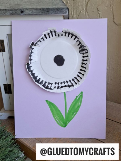 A craft paper plate dandelion with black marker lines for petals, a black dot center, and green marker stem and leaves, displayed on a purple background. The handle @GLUEDTOMYCRAFTS is printed at the bottom.