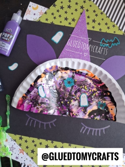 A paint splat unicorn craft comes to life on a paper plate, adorned with a whimsical purple hat, star stickers, ghost cutouts, and vibrant paint. Nearby, you’ll find a bottle of puffy paint, colorful paper scraps, and a playful green plastic skeleton. Text reads: @GLUEDTOMYCRAFTS.