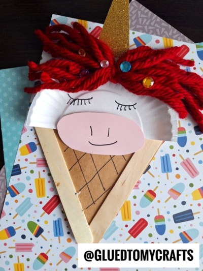 A unicorn ice cream cone craft made with a paper plate, red yarn for the mane, gold glitter paper for the horn, sequins, colored paper, and a popsicle stick unicorn accent, displayed on patterned sheets.