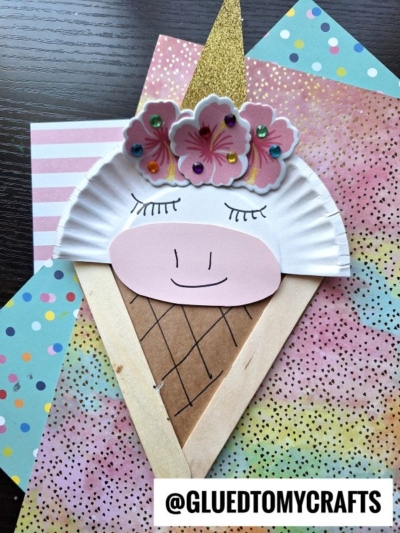A popsicle stick unicorn ice cream craft features a paper plate decorated with pink flowers, a golden horn, and a smiling face, set on colorful patterned papers. The watermark @GLUEDTOMYCRAFTS appears at the bottom.