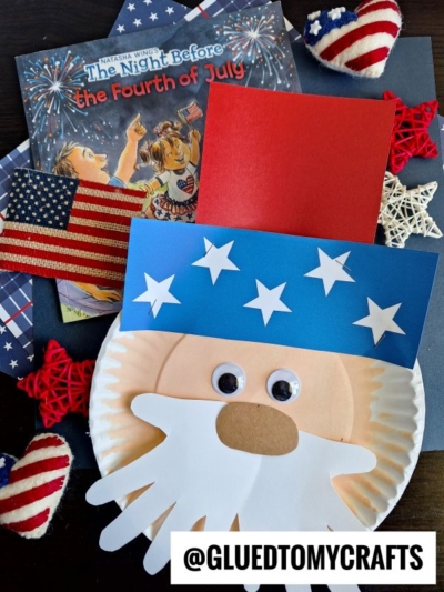 A paper plate Uncle Sam, complete with googly eyes and a paper Uncle Sam hat adorned with stars, rests on a Fourth of July-themed book amid patriotic decorations and an American flag. @GLUEDTOMYCRAFTS is written at the bottom.