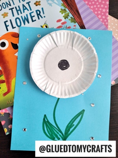 A handmade card features a white paper dandelion made from a cupcake liner on blue paper, with a drawn stem and leaves. Sparkling gems are scattered as accents. Patterned paper and a colorful book are in the background. Text reads @GLUEDTOMYCRAFTS.