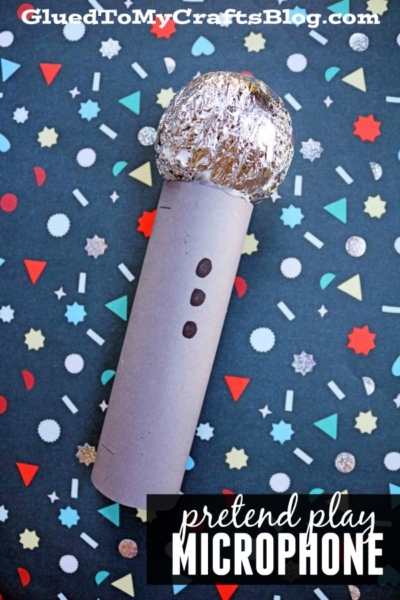 A homemade microphone craft for kids featuring a cardboard tube and a foil-covered ball, set on a colorful, patterned background sprinkled with confetti shapes.