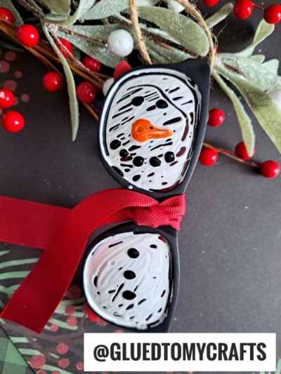 Handmade snowman ornament painted on black rocks, inspired by sunglasses turned snowman style, featuring an orange carrot nose, black eyes and mouth, red ribbon bowtie, with festive greenery and berries. @GLUEDTOMYCRAFTS at the bottom.