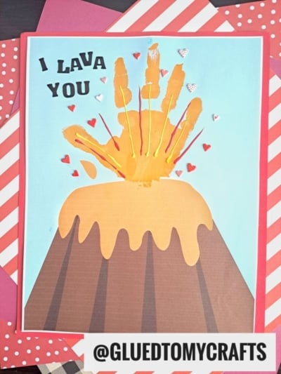 A handmade handprint volcano keepsake card shows a volcano erupting with a yellow handprint as lava, red and white heart sparks, and "I LAVA YOU" above, all bordered by red and white striped paper.