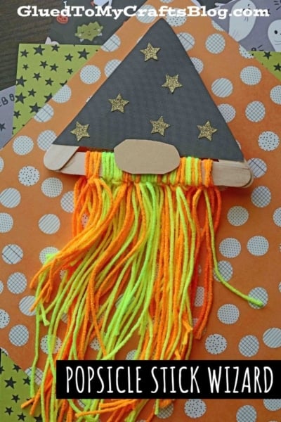 A craft wizard made with popsicle sticks, sporting a popsicle stick wizard hat crafted from black paper adorned with gold stars and finished off with a neon yarn beard, displayed on vibrant orange Halloween-themed paper.