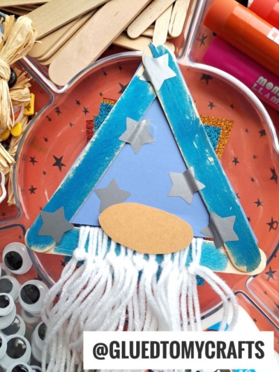 A blue popsicle stick wizard hat decorated with silver stars and white yarn resembling a beard sits on an orange tray with star patterns. Supplies like googly eyes, glue sticks, and raffia surround it.
