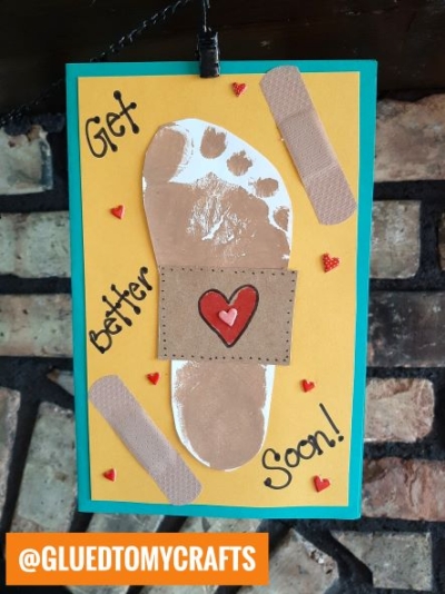 A handmade get well kid craft card features a tan Footprint Band-Aid with a paper bandage and red heart, surrounded by cut-out bandages, small red hearts, and the text Get better soon! on a bright yellow background.