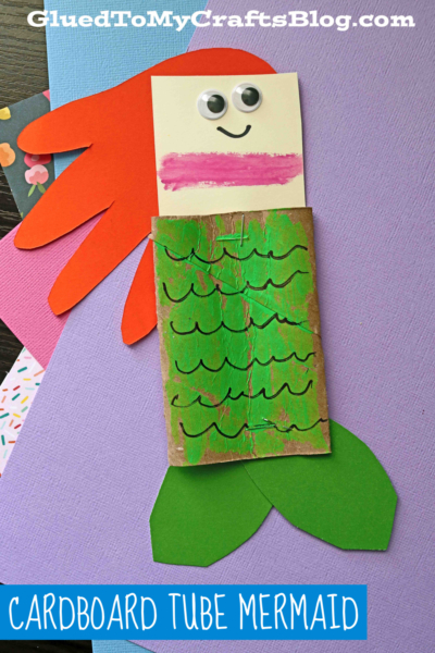 A handmade paper tube mermaid craft features a paper face with googly eyes, red hair, a painted cardboard tube body with green scales, and green paper fins on a purple background. Text reads CARDBOARD TUBE MERMAID.