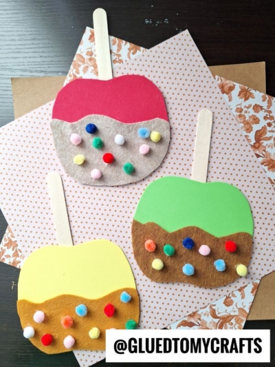 Three craft caramel apple friends, made from colorful felt and decorated with small pom-poms, sit on wooden popsicle sticks and patterned paper. The handle @GLUEDTOMYCRAFTS is visible at the bottom.
