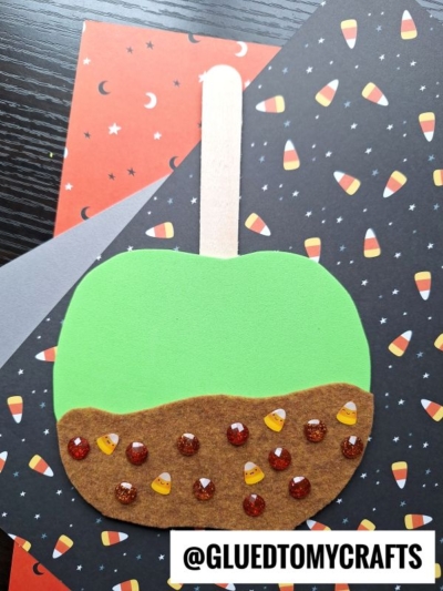 A paper craft of caramel apple friends with green and brown sections, decorated with glitter dots and small candy corn stickers, is placed on Halloween-themed paper. @GLUETOMYCRAFTS is labeled at the bottom.