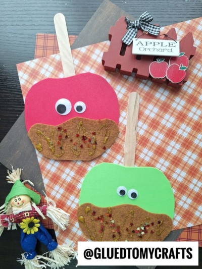 Two caramel apple friends with popsicle stick handles, googly eyes, and brown glitter tops are displayed on plaid paper beside a small scarecrow and a red Apple Orchard sign. @GLUEDTOMYCRAFTS is written at the bottom.