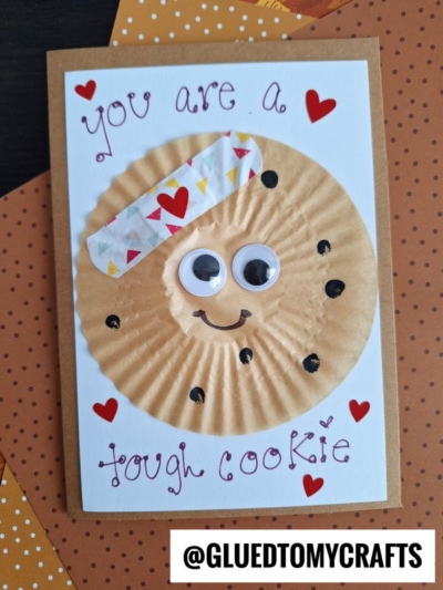Handmade you’re one tough cookie card featuring a googly-eyed cookie, a colorful bandage, hearts, and the uplifting message “You are a tough cookie.” The card is displayed on a charming brown polka dot background. Instagram handle @GLUEDTOMYCRAFTS is at the bottom.