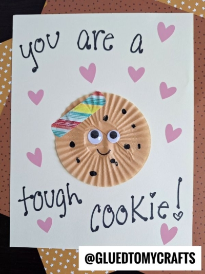A handmade “you are one tough cookie card” features a cookie crafted from a cupcake liner, googly eyes, a colorful paper beret, and drawn chocolate chips. Pink hearts surround the cookie. @GLUEDTOMYCRAFTS is printed at the bottom.