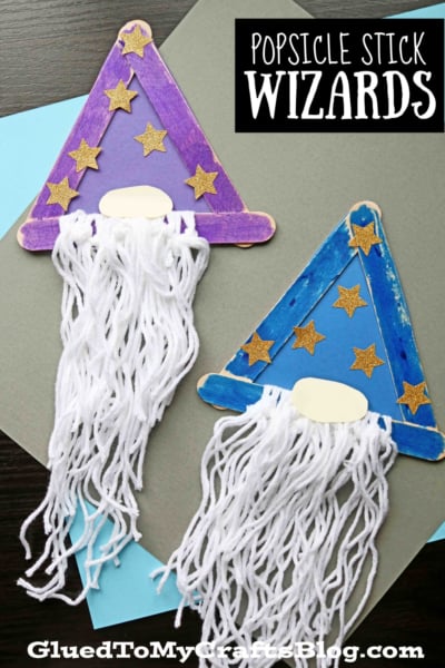 Two popsicle stick crafts shaped like wizards with triangle popsicle stick wizard hats, gold star decorations, white yarn beards, and oval noses. One hat is purple, the other blue, displayed on colored paper. Text reads Popsicle Stick Wizards.