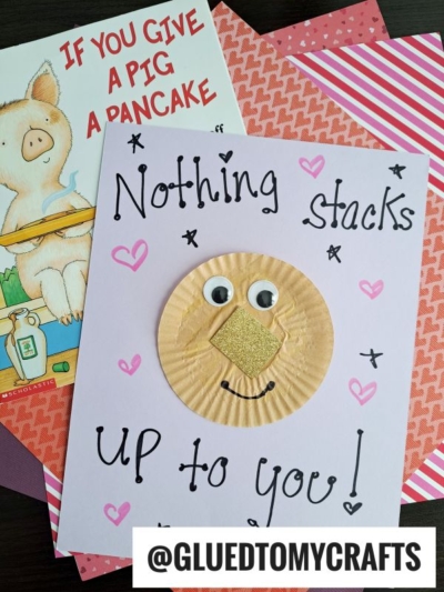 A handmade card with “Nothing stacks up to you!” written in black marker, decorated with hearts, stars, and a pig face made from a cupcake liner pancake, googly eyes, a gold glitter snout, and a curved mouth. A pig book is nearby.