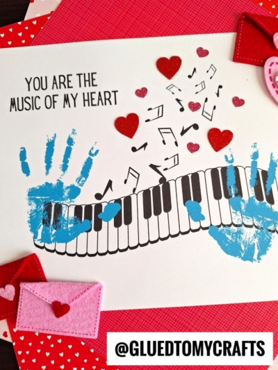 A handmade card with blue handprints, a piano keyboard, musical notes, red heart stickers, felt hearts, and envelopes—perfect for celebrating National Grandparent’s Day. Text reads: You are the music of my heart. @GLUEDTOMYCRAFTS is at the bottom.