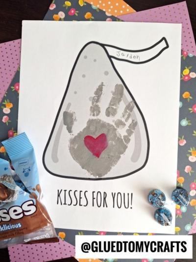 A children’s craft features a grey Hershey’s Kiss outline with a handprint and red heart in the center—a sweet kisses keepsake! "KISSES FOR YOU!" is written below, with Jayden on the tag. Chocolate candies and colorful paper surround it.