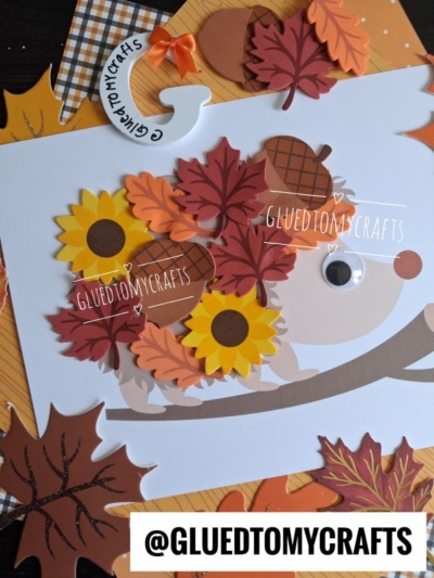 A handmade fall card features a charming leaf hedgehog decorated with paper sunflowers, acorns, and autumn leaves. The background shows more fall-themed paper, and the text @GLUEDTOMYCRAFTS is displayed at the bottom.