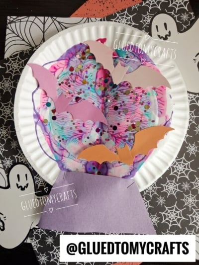 A Halloween craft made from a paper plate and construction paper, featuring bats, ghosts, and paint splat swirls, on a spiderweb-patterned background. Text reads @GLUEDTOMYCRAFTS.