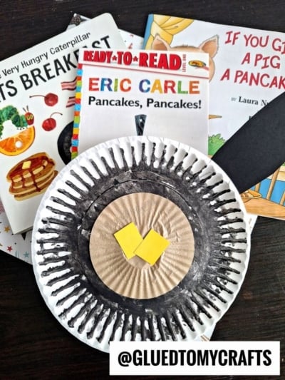 A paper plate craft, featuring a cupcake liner pancake with syrup and butter, sits atop three children’s books about pancakes, including Pancakes, Pancakes! by Eric Carle. A black strip and handle next to the craft resemble a spatula.