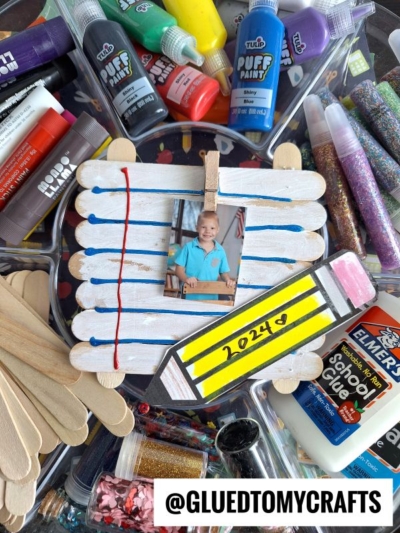 Popsicle stick photo frame decorated with a painted pencil labeled 2024 and accented with popsicle stick notebook paper, holding a child’s school photo, surrounded by art supplies like paint, glue, glitter, and craft sticks.