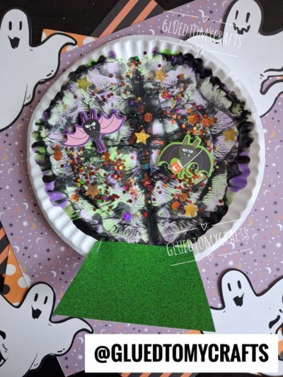 A Halloween craft of a spooky tree on a paper plate, featuring a paint splat background in black and purple, bat and cat stickers, sequins, and ghost decorations, set on a festive backdrop reminiscent of a witch’s ball. @GLUEDTOMYCRAFTS appears at the bottom.