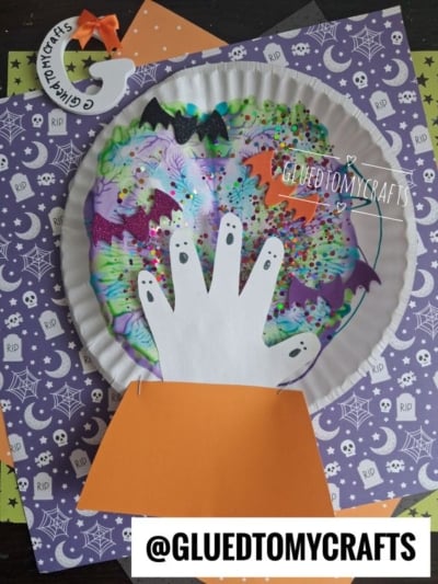 A Halloween craft featuring a paint splat witch’s ball made from a paper plate crystal ball, a white paper hand, glitter, bats, and stars, all displayed on festive spiderweb-themed paper. An @GLUEDTOMYCRAFTS label is at the bottom.