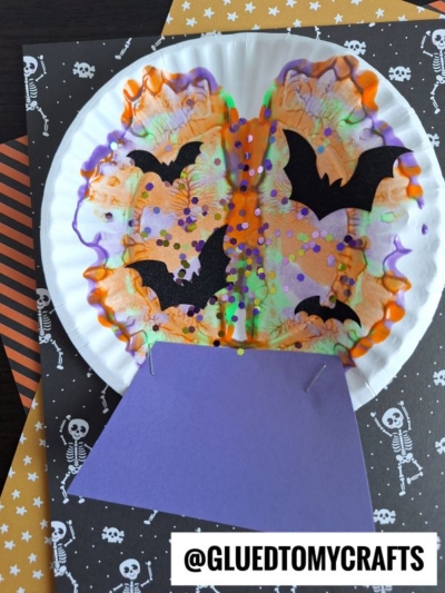 A paper plate craft shows a colorful cauldron with orange, purple, and green paint splat designs, black bats, and confetti. The plate sits on skeleton and striped paper. A purple paper triangle forms the base.